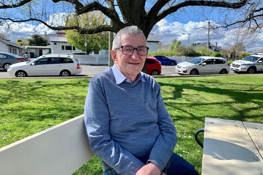 A 79-year-old man wearing a blue jumper and black glasses sits with arms crossed in the sun at a picnic table in the park.
