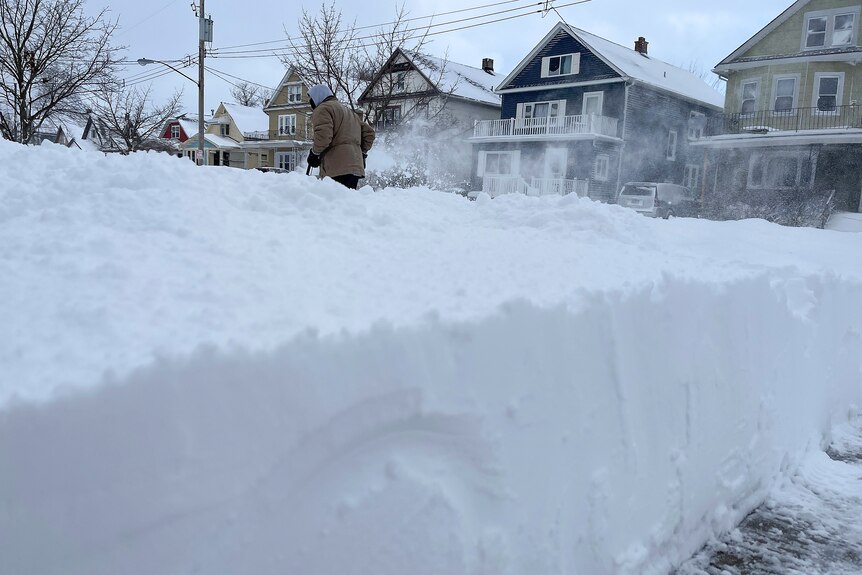 A very thick layer of snow is pictured, behind it some houses and someone trying to remove the snow.
