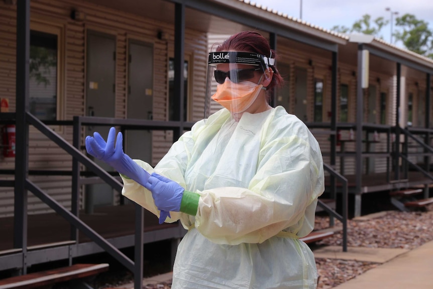 Un trabajador de la salud con EPP completo usa guantes afuera cerca de algunos Dungas en las instalaciones de cuarentena de Howard Springs.