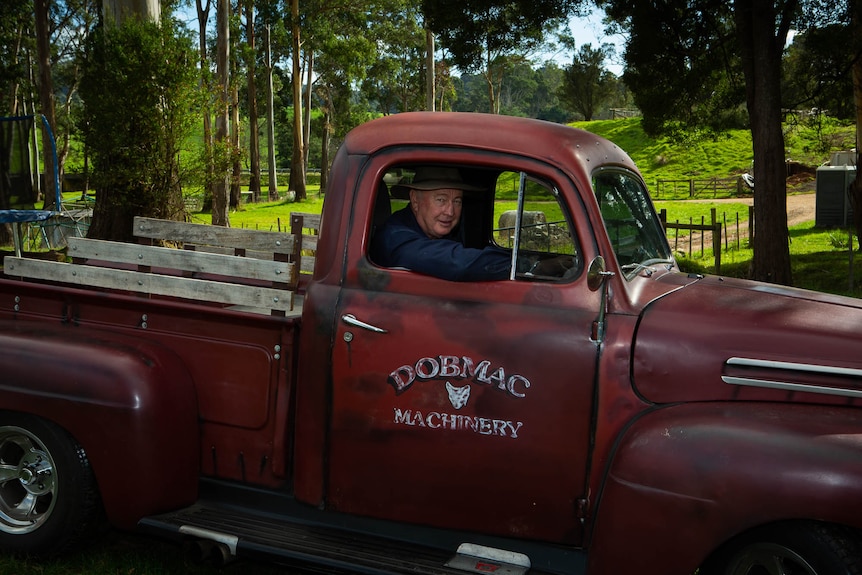 A man at the wheel of an old red pickup truck, smiling out the driver side window.