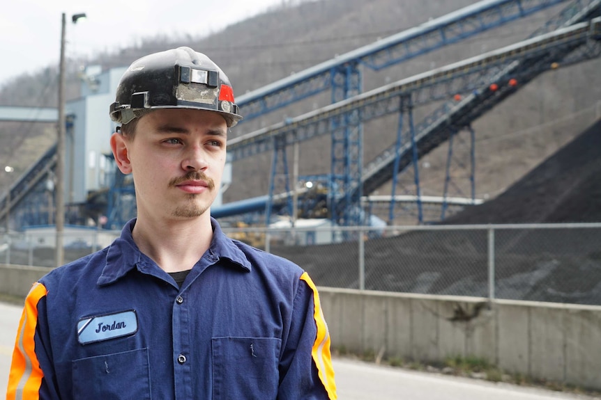 A young man with facial hair wears a hard hat and mining wear at a coal mining site in west virginia.