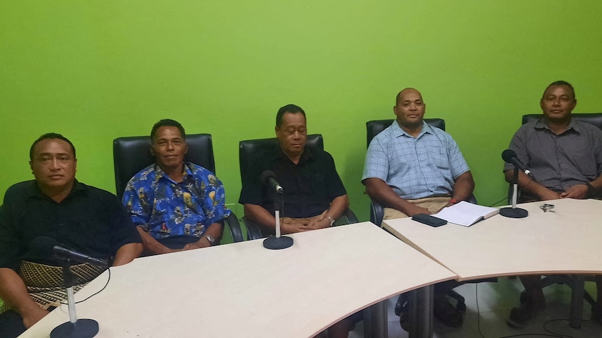 Four men sit in front of a green wall and desk