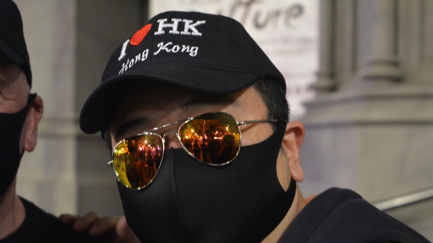 A man in a face-mask, aviator sunglasses and a cap that reads 'I <3 HK' looks directly into the camera.