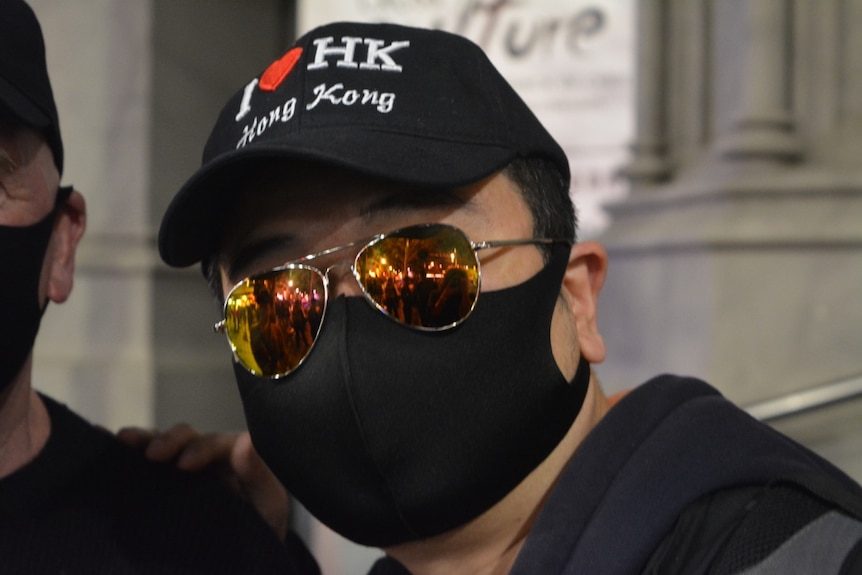 A man in a face-mask, aviator sunglasses and a cap that reads 'I <3 HK' looks directly into the camera.