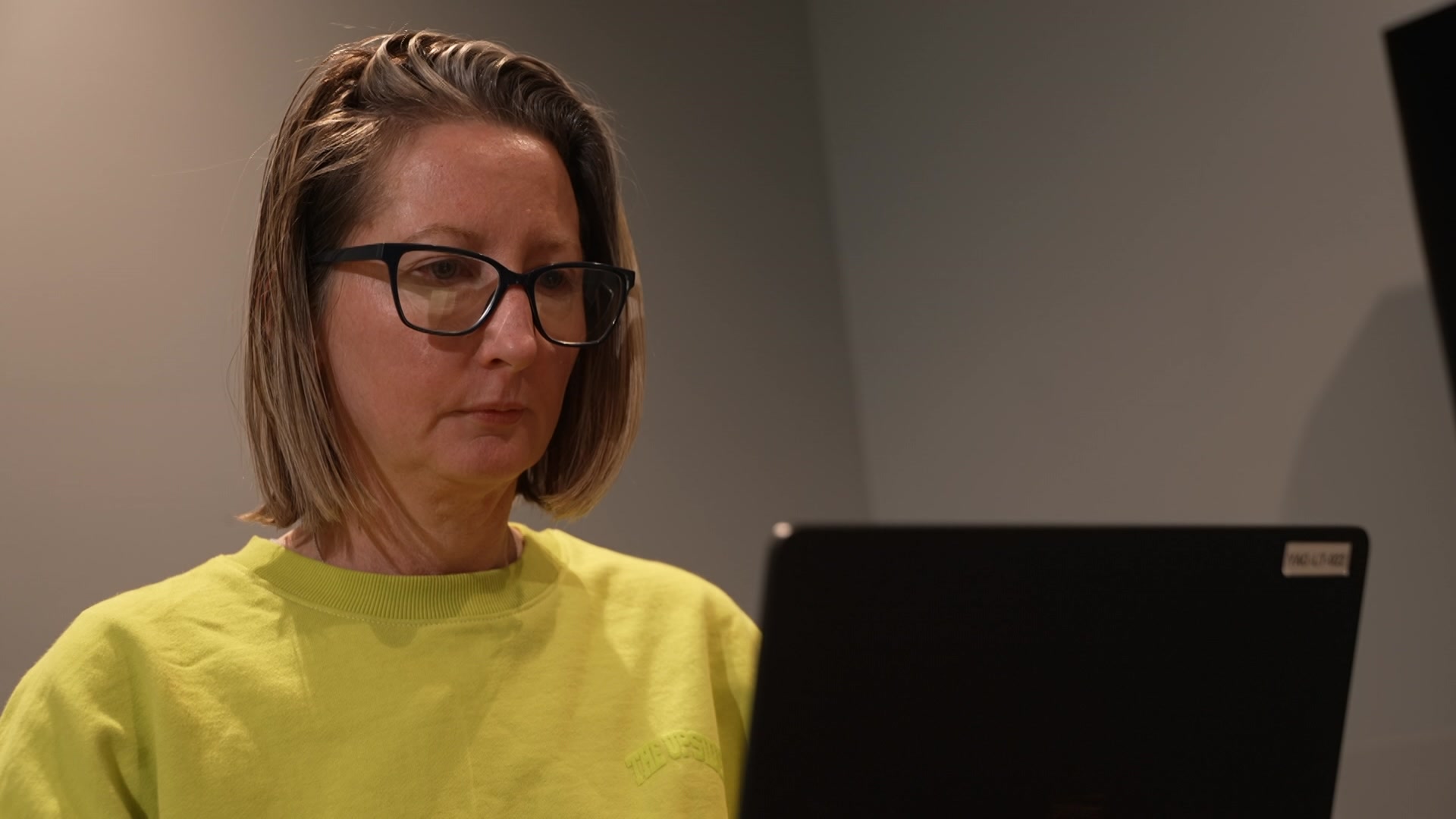 Una mujer con camisa amarilla y gafas mira la pantalla de la computadora.