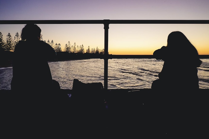 A mother and daughter sit on a pier at sunrise looking at the water