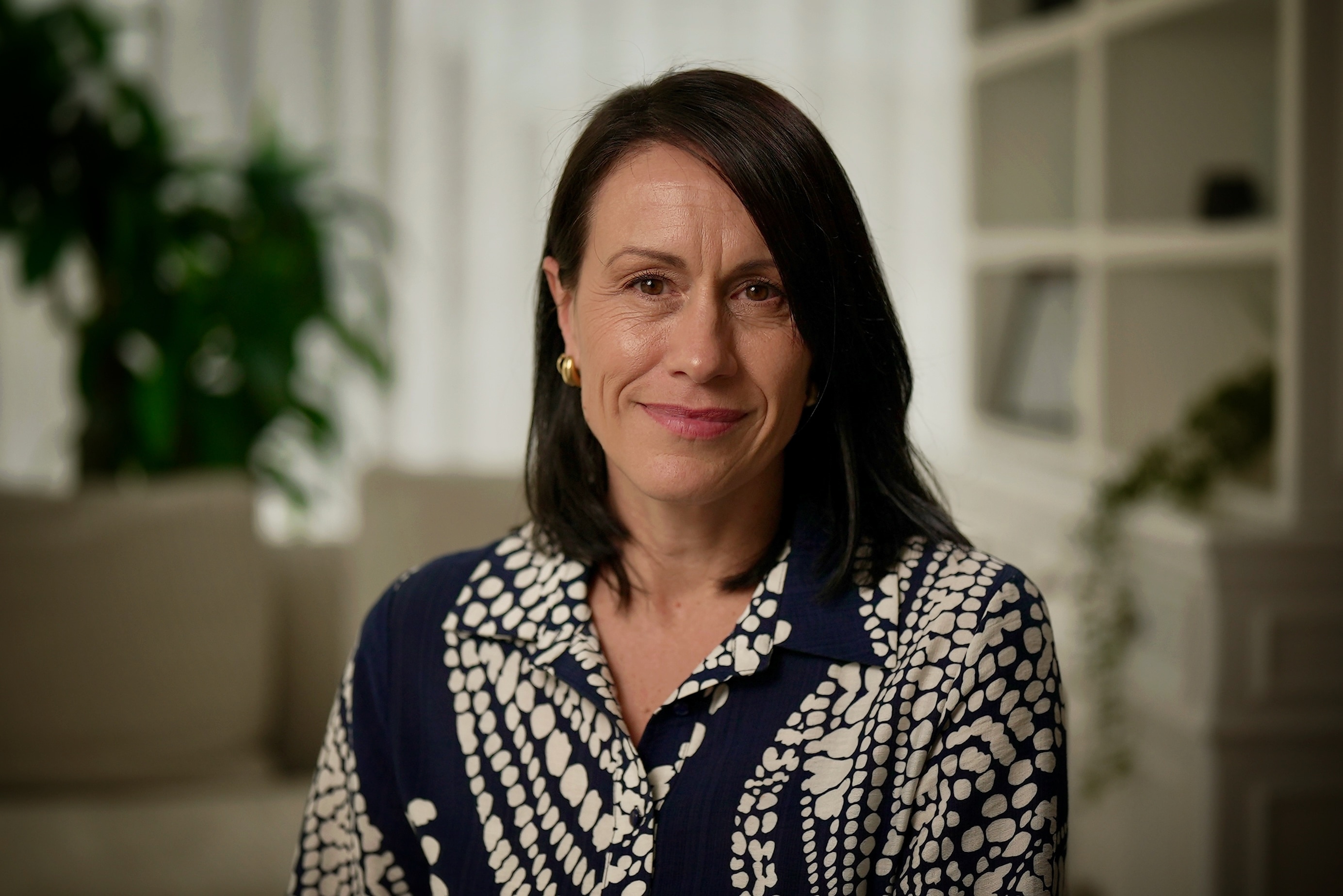 Dark-haired woman wearing professional clothing sits on a sofa and looks at the camera with a small smile.