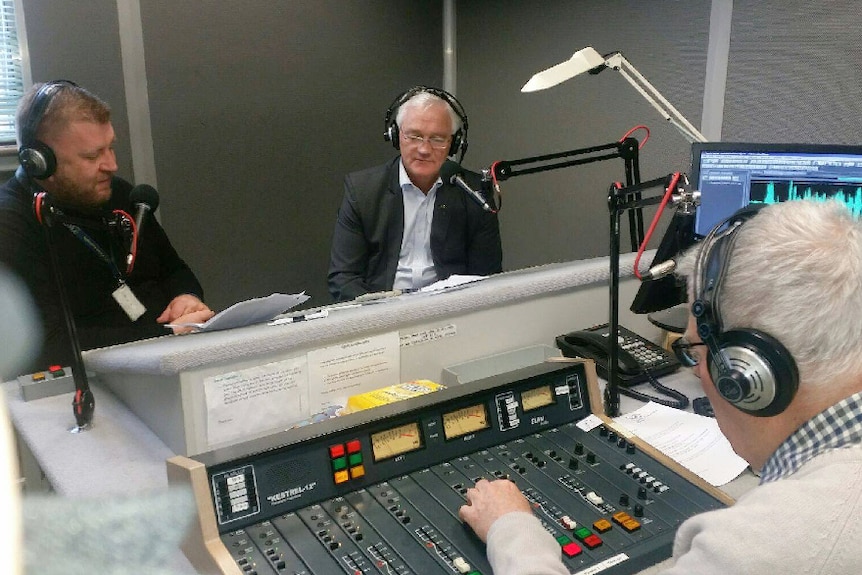 Three men sitting in a radio studio with headphones on and looking at papers in front of them.