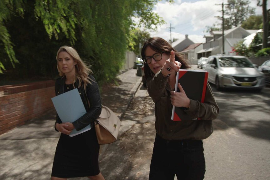 Worthington and Meldrum-Hanna walking in a street with notebooks in arms and Meldrum-Hanna pointing ahead.