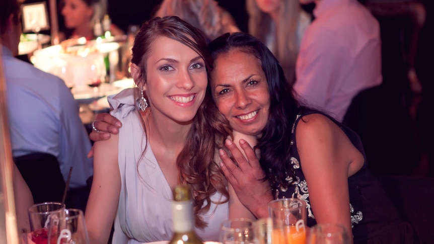 Apryl, wearing a formal dress, smiles as Tanya Day smiling, rests her head on her shoulder, at a formal dinner setting