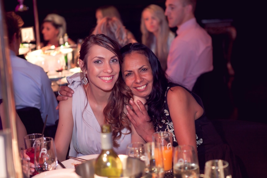 Apryl, wearing a formal dress, smiles as Tanya Day smiling, rests her head on her shoulder, at a formal dinner setting