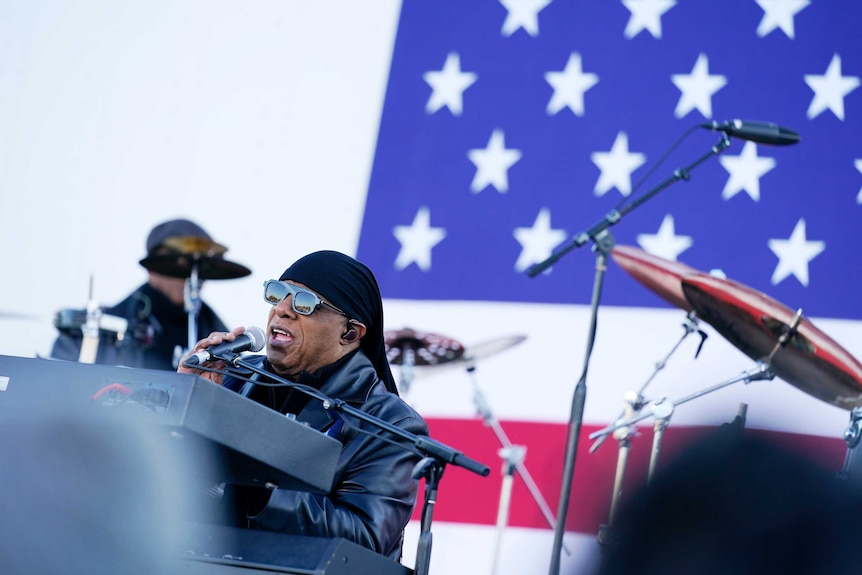 Stevie Wonder, wearing a bandana and sunglasses, sings while playing a keyboard in front of an American flag.