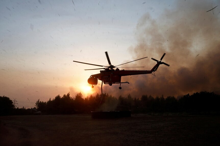 A silhouetted helicopter flies above the tree line at dusk with a fire burning behind it in the distance