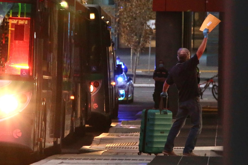 A man with a suitcase and his arm in the air waving an envelope walks away from a bus at dusk towards a hotel.