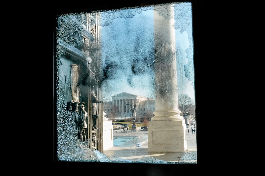 A burned and broken window gives a glimpse of the US Supreme Court building under a blue sky