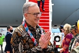 Anthony Albanese, wearing a batik top, holds his hands in the pray position while greeting Indonesian officials at the airport