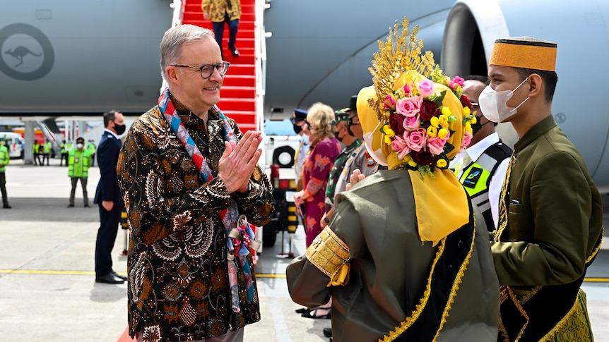 Anthony Albanese, wearing a batik top, holds his hands in the pray position while greeting Indonesian officials at the airport