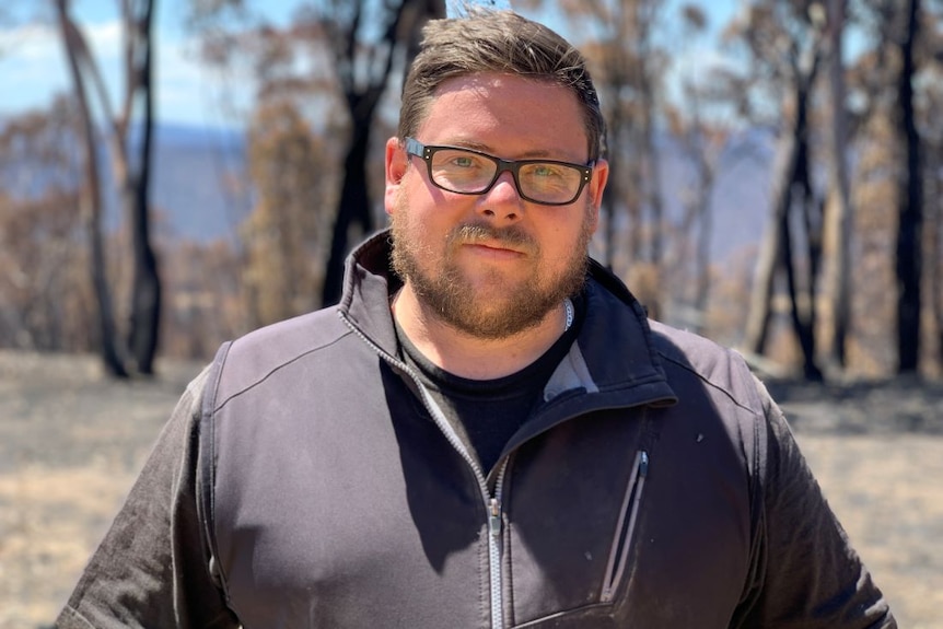 A man with black glasses stands in front of gum trees.