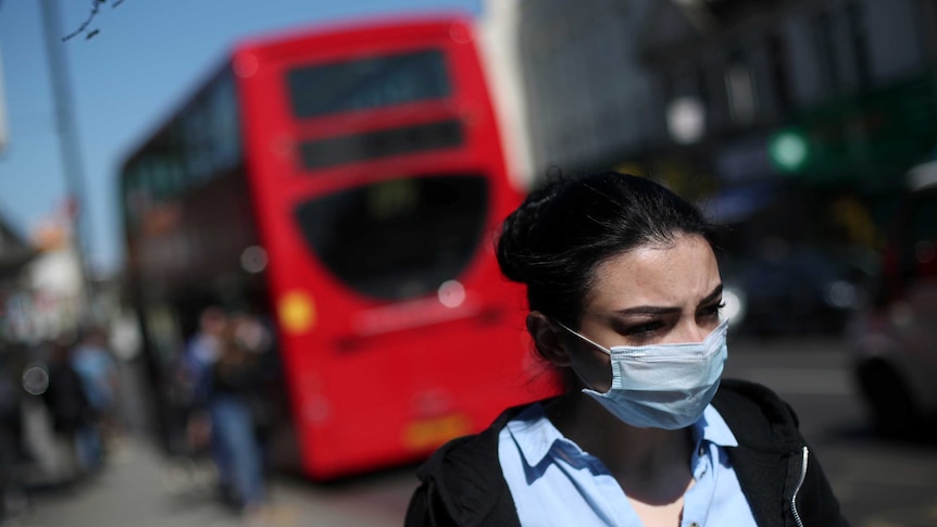A woman wearing a mask walks down a street with a London bus blurred in the background