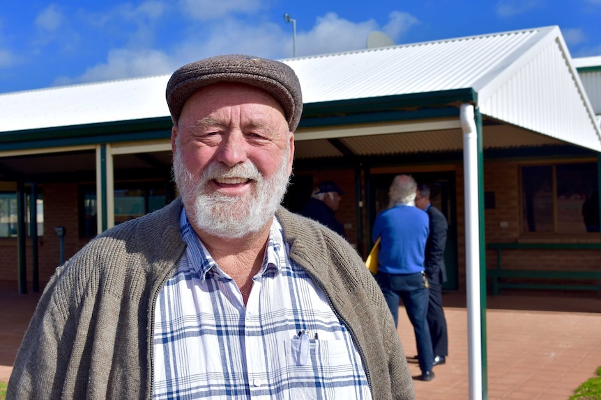 A man with a white beard stares at the camera.  He is wearing a cap and the sky is blue.