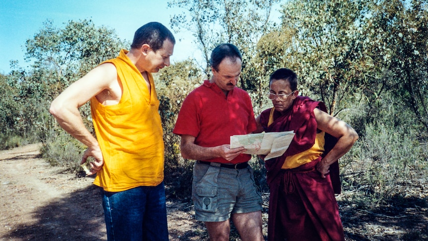 Ian holding a map flanked by two people, one a monk in robes.
