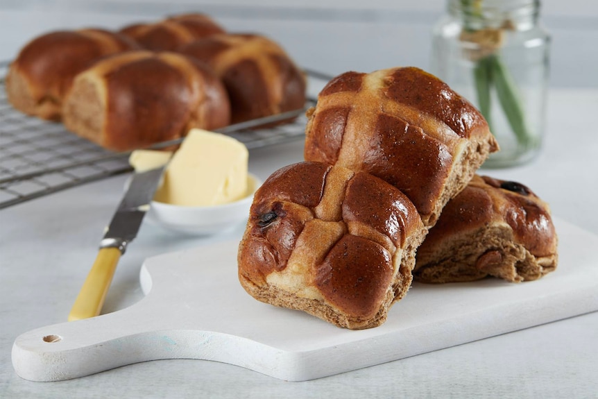 A few hotcross buns on a small plate with a butter knife in butter behind it and a daffodil in a vase.