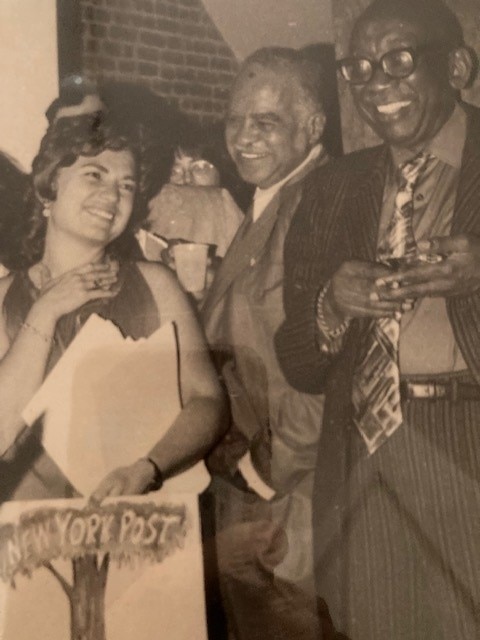 A black and white photo of a woman with cropped dark hair standing, smiling surrounded by men