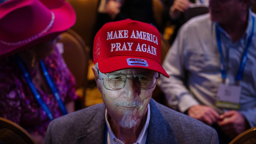 An elderly man with glasses and a white beard where's a red cap with the words "make america pray again"