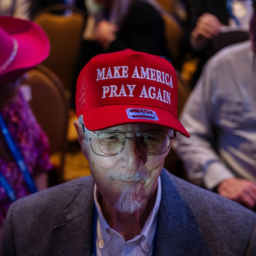 An elderly man with glasses and a white beard where's a red cap with the words "make america pray again"