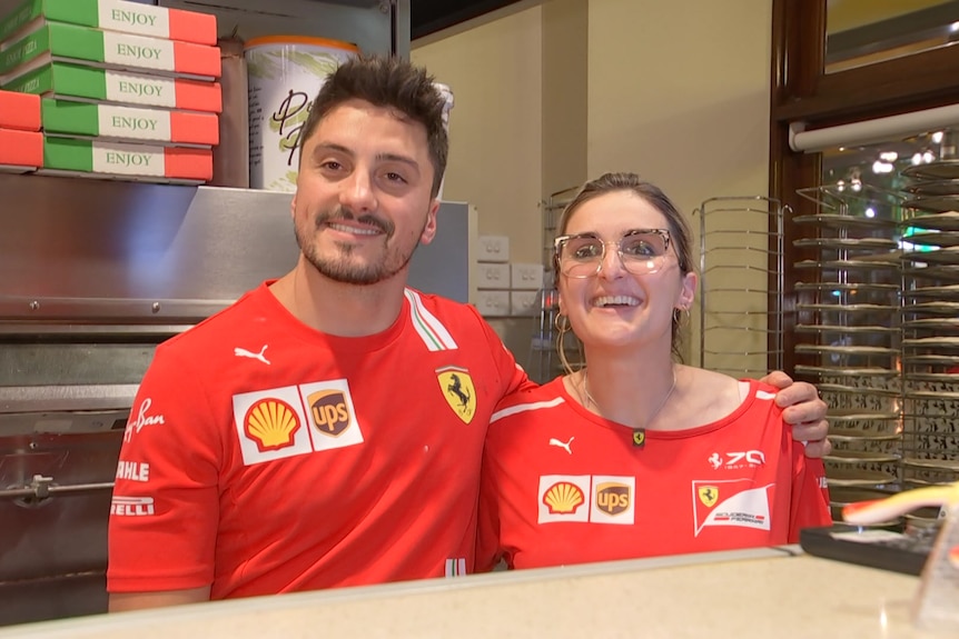 A man and woman wearing red Ferrari formula one shirts stand behind the counter of a pizza restaurant.