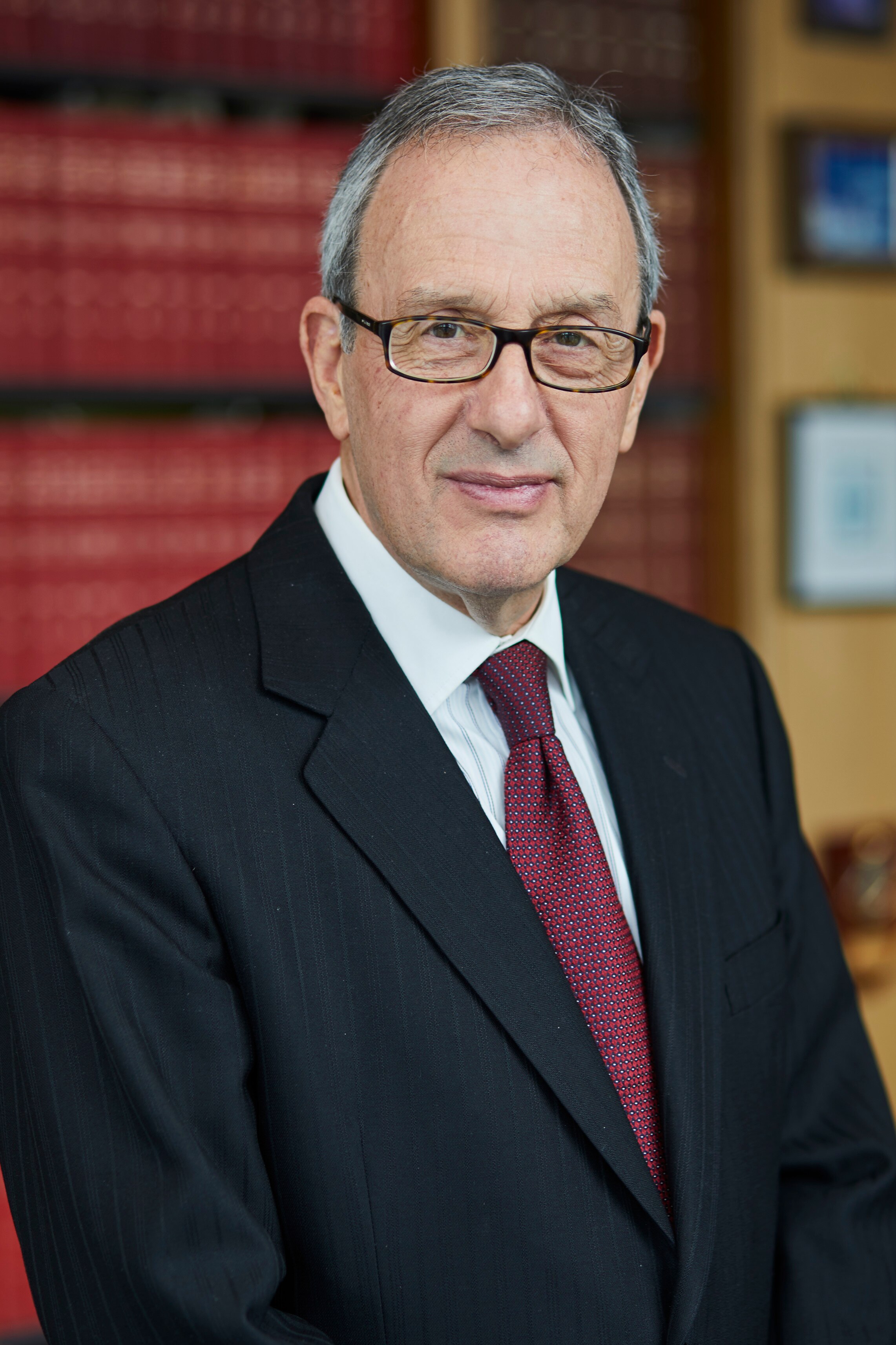 An older man wears glasses and stands in front of a bookcase full of books