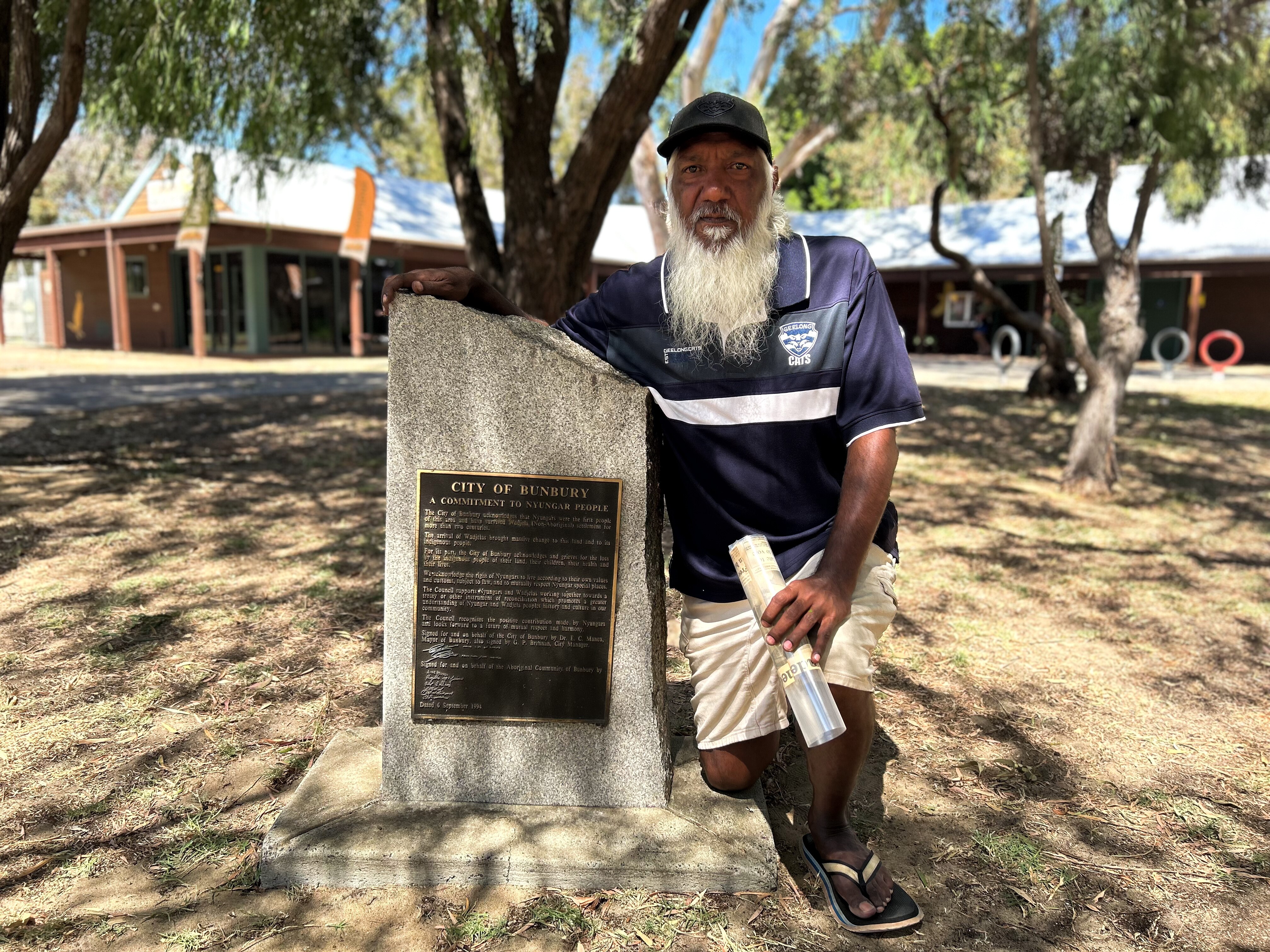 An indigenous man with a white beard and black cap kneels next to a plaque mounted on a stone in a park