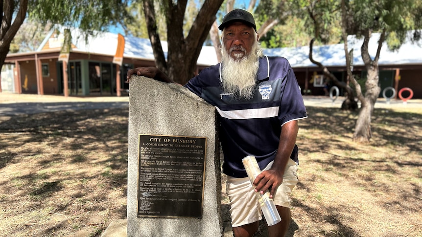 An indigenous man with a white beard and black cap kneels next to a plaque mounted on a stone in a park