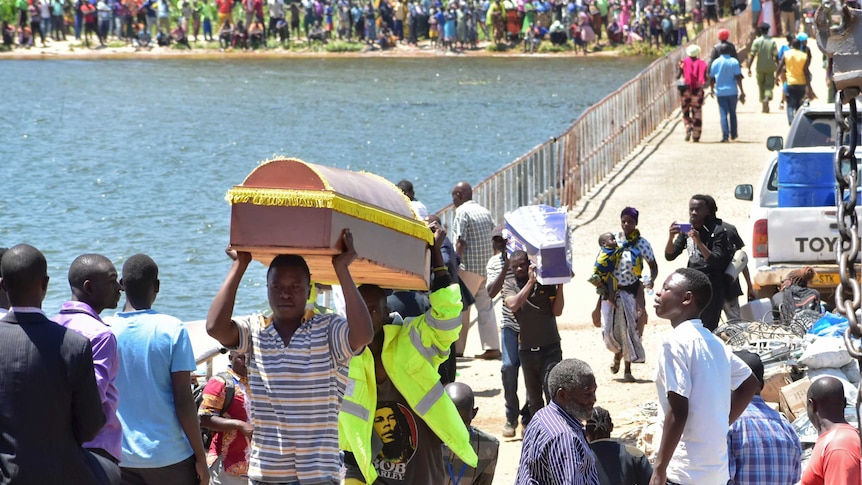 Men carry coffins along a crowded wharf with many people waiting on the shores in the background