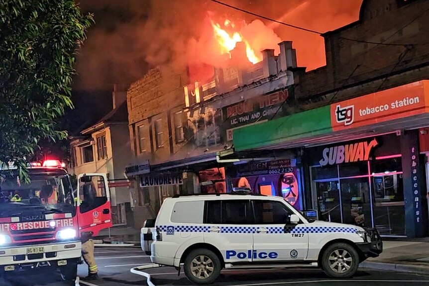 A fire truck and police car parked in front of a shop with tall flame above the roof.