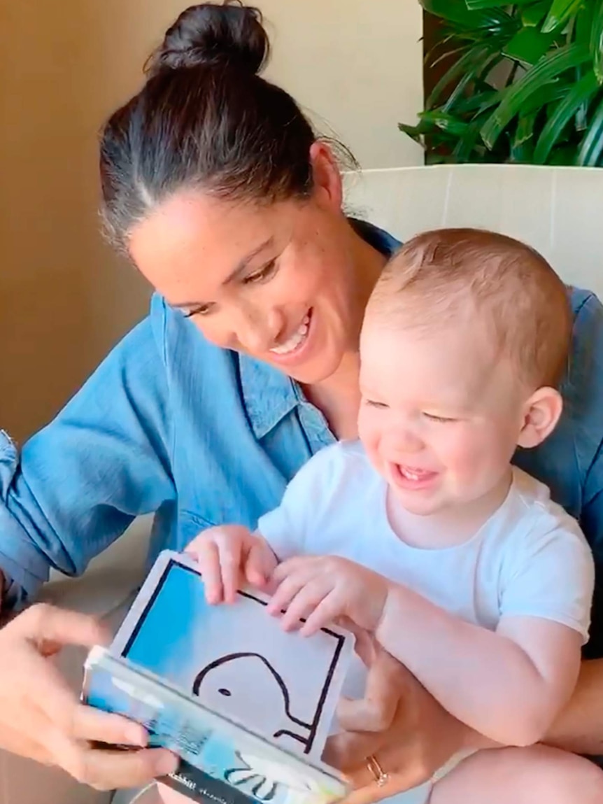 Woman with dark hair tied back in a bun wearing blue collared dress with one-year-old boy with blonde hair, white shirt, reading