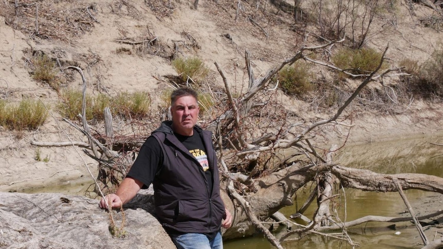 Graeme McCrabb leaning against a tree and standing in a shallow pool of water along the Darling River