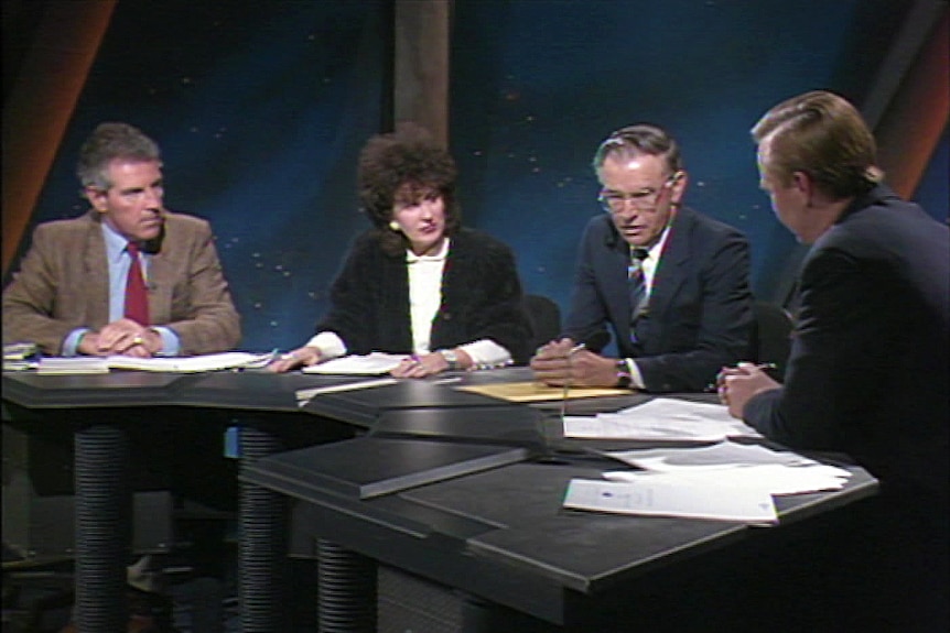 A historical image of four people sitting around a desk, one is a male reporter interviewing a woman and two other men.