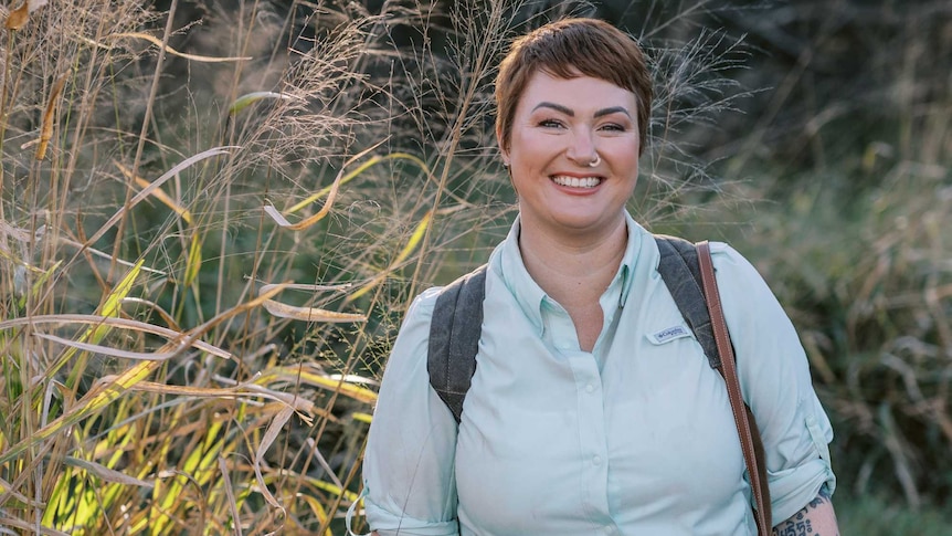 Woman in hiking gear with backpack surrounded by long grass and trees smiling into camera.