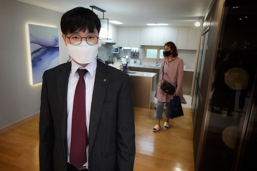 A young Korean man in a face mask and a suit stands in a kitchen while a woman walks past him 