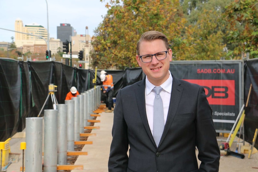 A man in a suit stares at the camera, while workmen install bollards behind him.