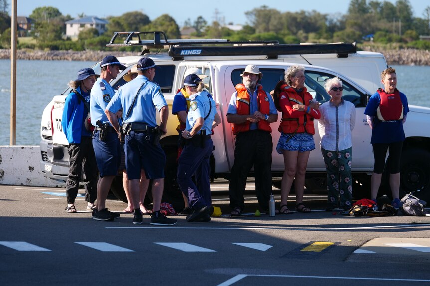 group of people in lifejackets line up on ute as police question them. 