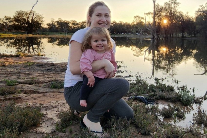 A woman is crouching holding a young child standing by a river. They are smiling.