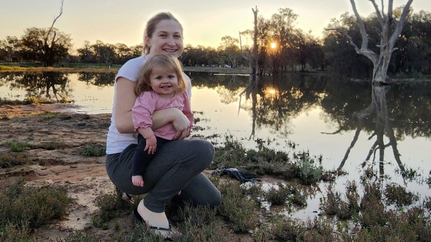 A woman is crouching holding a young child standing by a river. They are smiling.