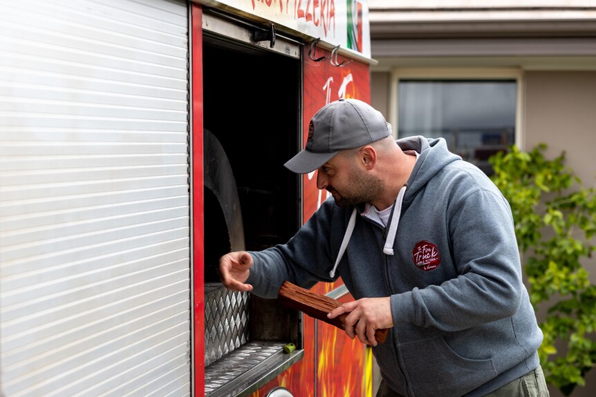 A man in a cap and hoodie putting wood into an oven inside a red truck.