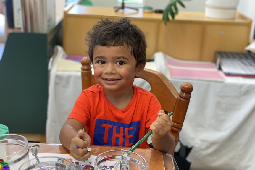 A little boy smiles as he plays with a paintbrush. He is sitting at a table