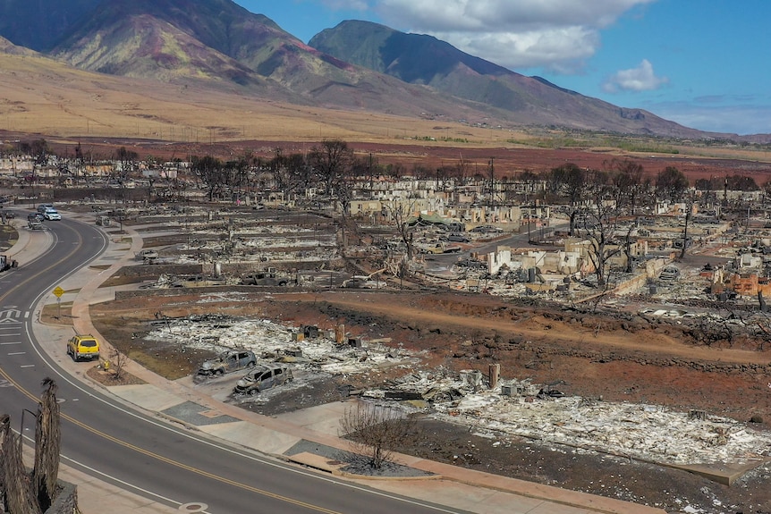 A yellow car drives around a bend in a road next to the burnt-out remains of buildings