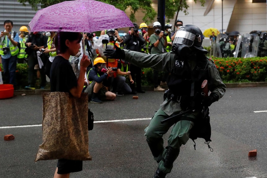 A woman with a pink umbrella stands calmly in front of a police office with threatening body language.