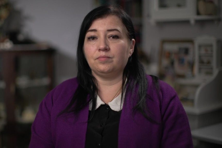 A woman sits indoors, looking ahead. Her eyes have tears.