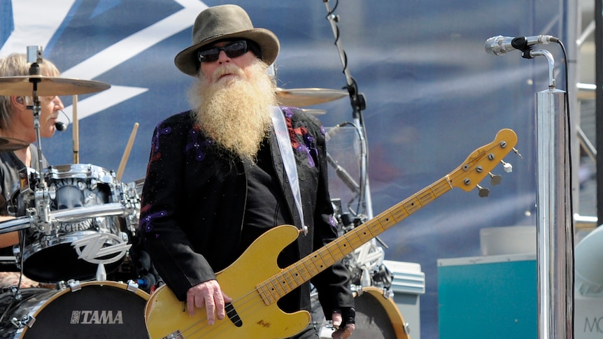 Dusty Hill onstage at a daytime concert, holding a bass guitar, with the iconic long beard, sunglasses and hat. 